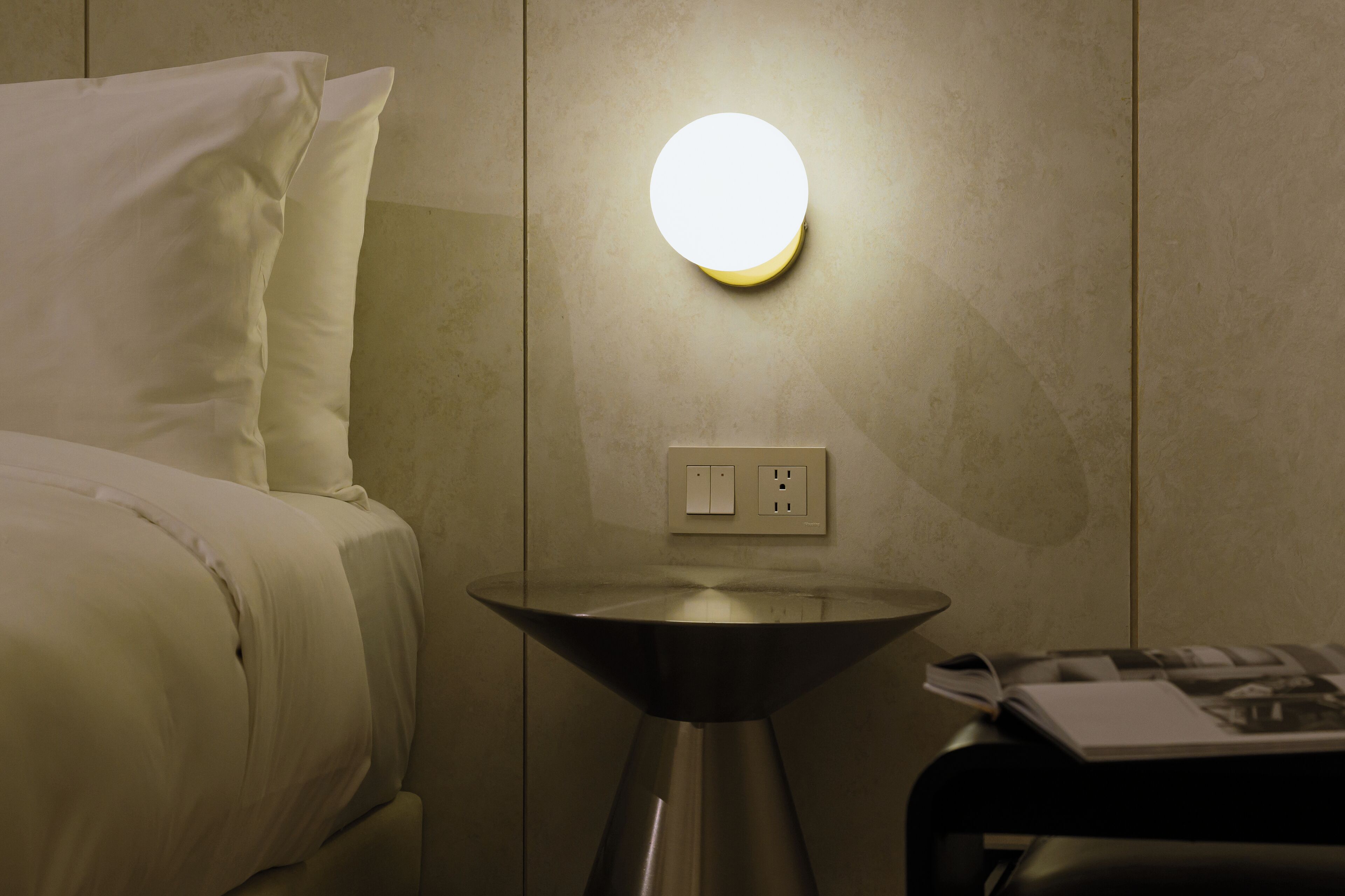 En pænt redt seng med hvidt sengetøj, et lille rundt bord med metalstel og en vægmonteret lampe.