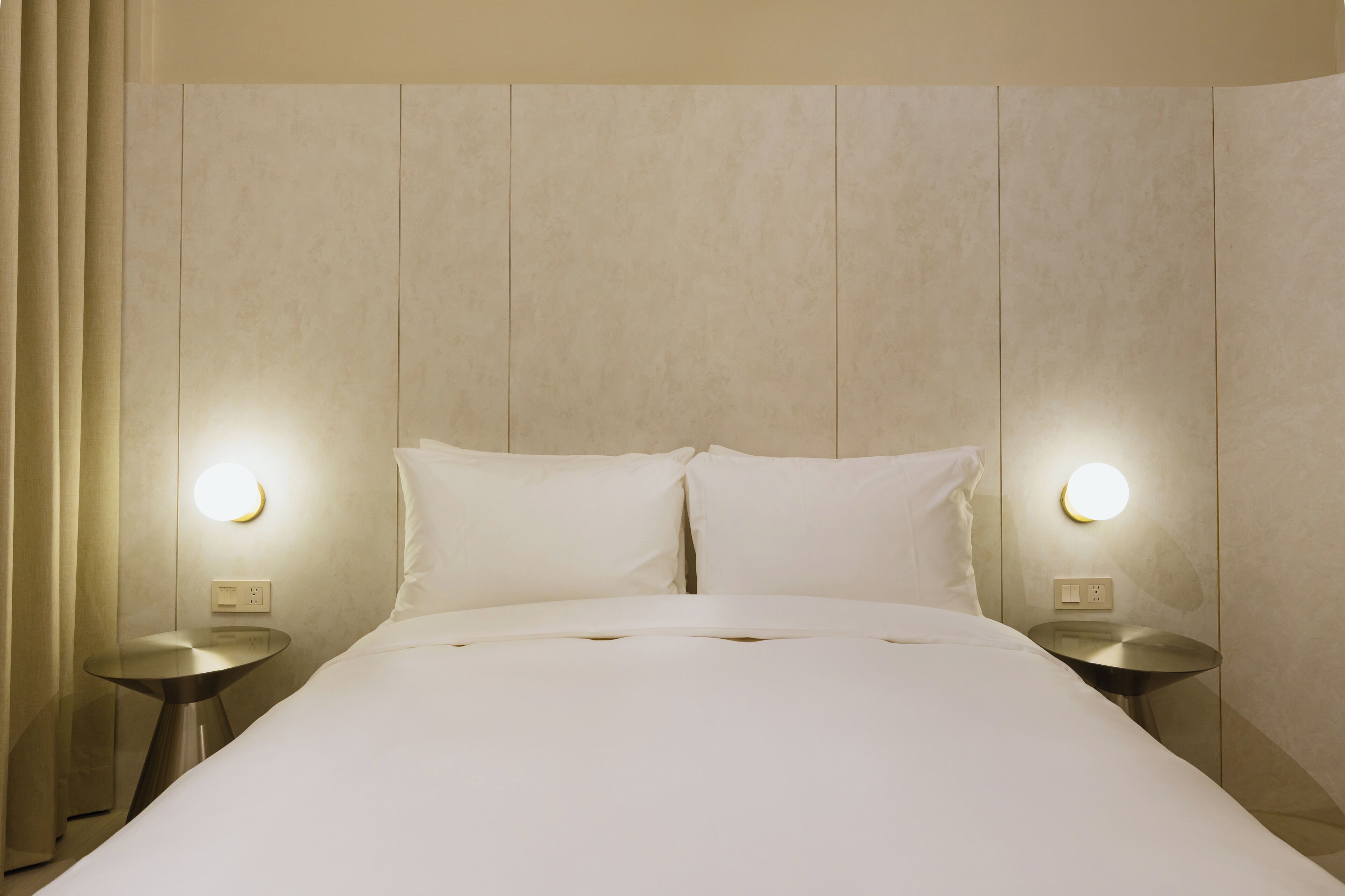 Una cama bien hecha con dos almohadas, una mesita redonda y un aplique de pared.