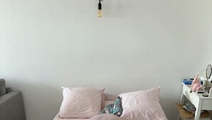 En snyggt bäddad säng med ett rosa överkast och kuddar, en vit byrå med en spegel och en väggmonterad lampa.