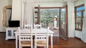 Una habitación con piso de madera, una mesa blanca con sillas, un televisor y un balcón con vistas a áreas verdes.