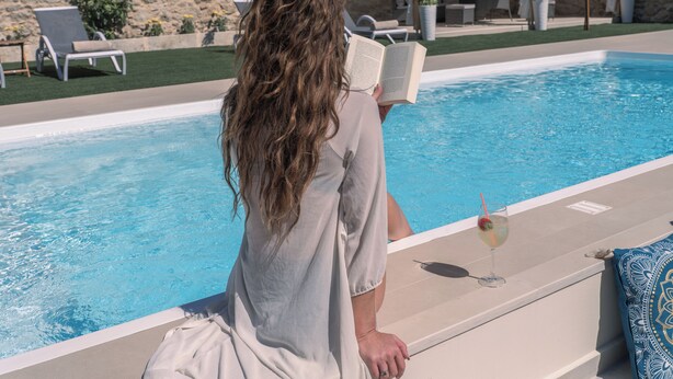 Una persona leyendo un libro junto a una piscina, con una bebida.