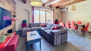A modern living room with a sectional sofa, a coffee table, and a TV mounted on a wooden wall. There is a dining area with red chairs and a table, and a large window providing natural light. - Mia - Your Gozo Haven with Pool (Ix-Xagħra)