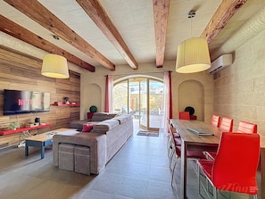 Un salon moderne avec un canapé, un téléviseur, des poutres en bois et un coin repas avec des chaises rouges.