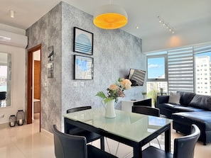 A modern living room with a glass dining table, black leather sofa, and a cityscape view through large windows. - 1BR apart, A/C, Wifi, Smart tv*Piantini-YellowKey (Santo Domingo)
