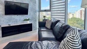 Un salón moderno con un sofá de cuero negro, un televisor montado en la pared y vistas a la ciudad a través de amplios ventanales.