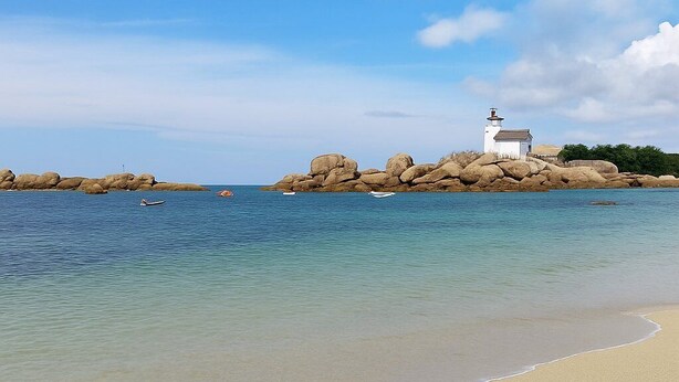 Une plage à l’eau claire, un bâtiment blanc surmonté d’un phare et de gros rochers.