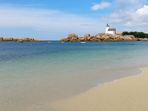 Une plage à l’eau claire, un bâtiment blanc surmonté d’un phare et de gros rochers.