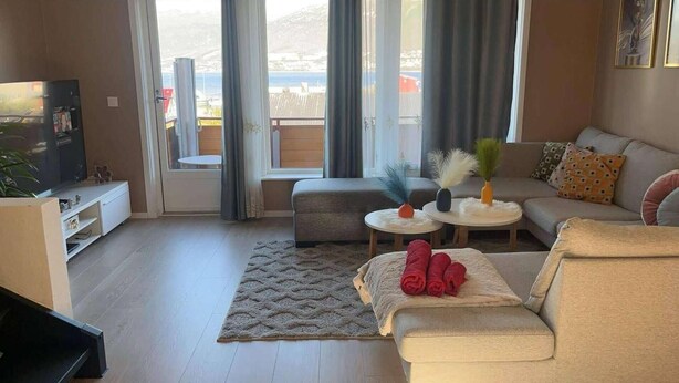 A modern living room with a sofa, a coffee table, and a TV. There are two windows with curtains, a view of the outdoors, and a small balcony. - Polar View Apartment (Tromsø)