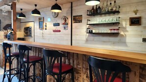 Eine Bar mit Holzwänden, schwarzen Barhocker, Pendelleuchten und Regalen mit Flaschen und Gläsern.