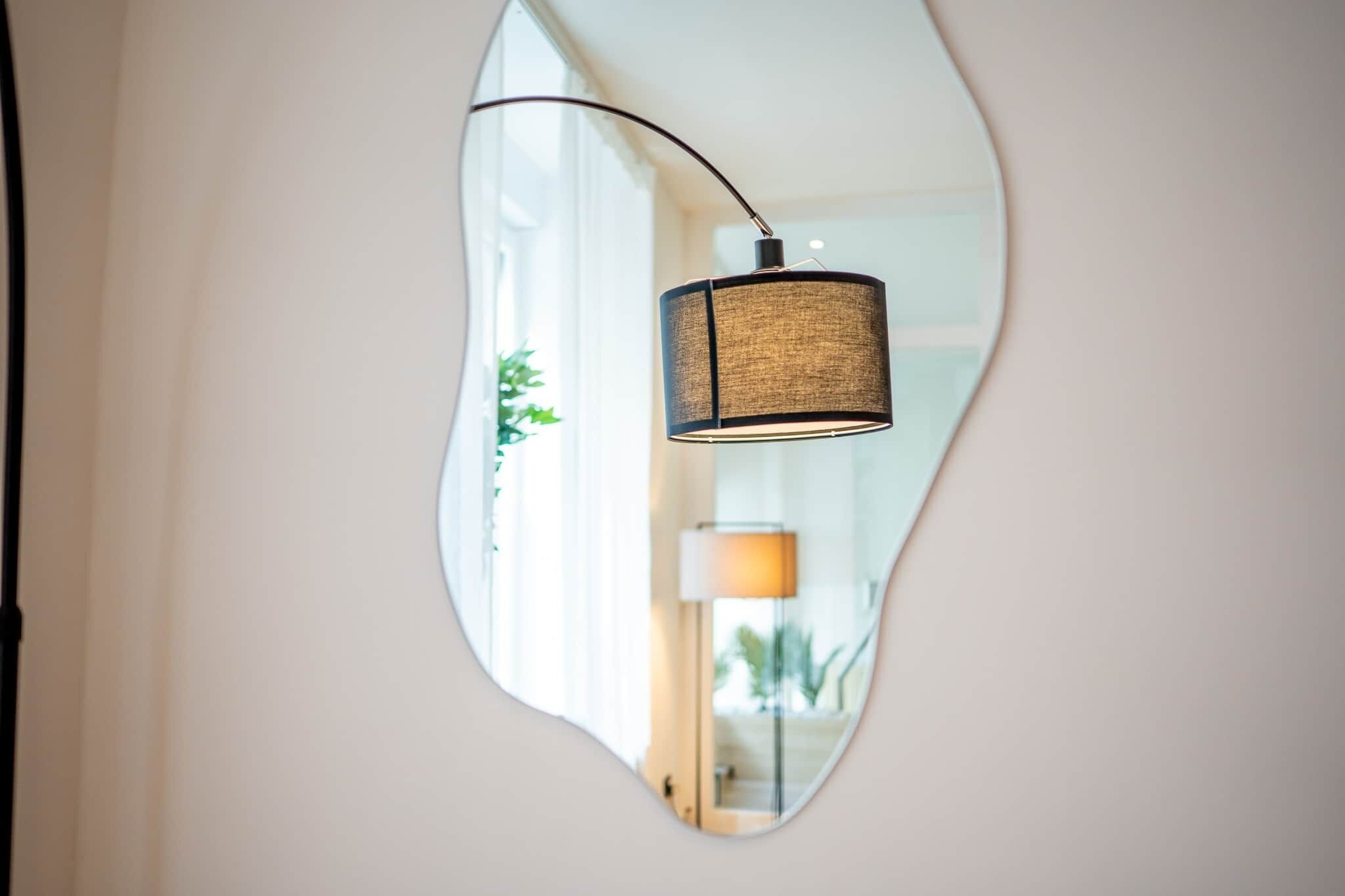 Een spiegel die een lamp en een kamer met een raam en een plantje weerspiegelt.