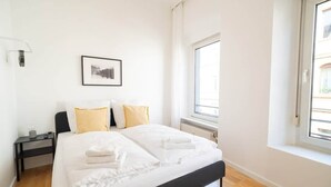 A neatly made bed with white linens and two pillows, a bedside table with a lamp, a framed picture on the wall, and a window with a view of buildings. - Holiday apartment | 2 bedrooms | Sauna | Terrace (Koblenz)