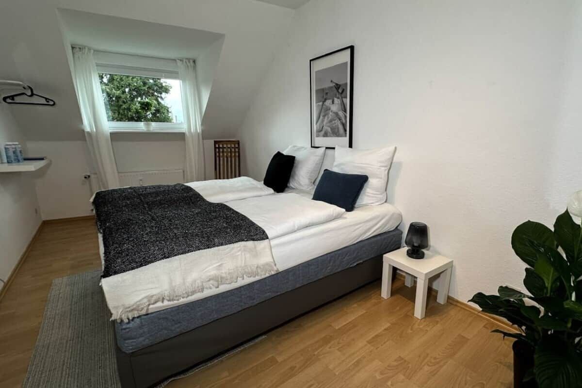A bedroom with a bed, a bedside table, a window with curtains, a framed picture on the wall, and a plant.