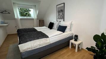 A bedroom with a bed, a bedside table, a window with curtains, a framed picture on the wall, and a plant.