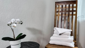 Una orquídea blanca en una maceta blanca sobre una mesa redonda negra, una silla de madera con toallas blancas dobladas y un cabecero de madera con listones.