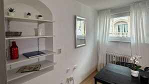 Una habitación con paredes blancas, una mesa negra, un espejo, un ventanal con cortinas y estantes con plantas y una botella roja.