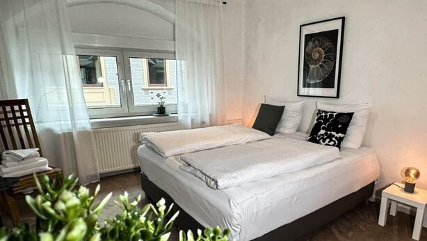 Un dormitorio con una cama grande, una ventana con cortinas, un radiador, una mesita con lámpara y un cuadro en la pared.