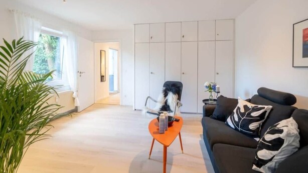 Un salon moderne avec un canapé, une table basse et un fauteuil. Au fond, on aperçoit une grande fenêtre avec des rideaux, une plante et une armoire.