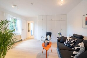 Una sala de estar moderna con un sofá, una mesa de centro y una silla. Se ve una ventana grande con cortinas, una planta y un armario al fondo.