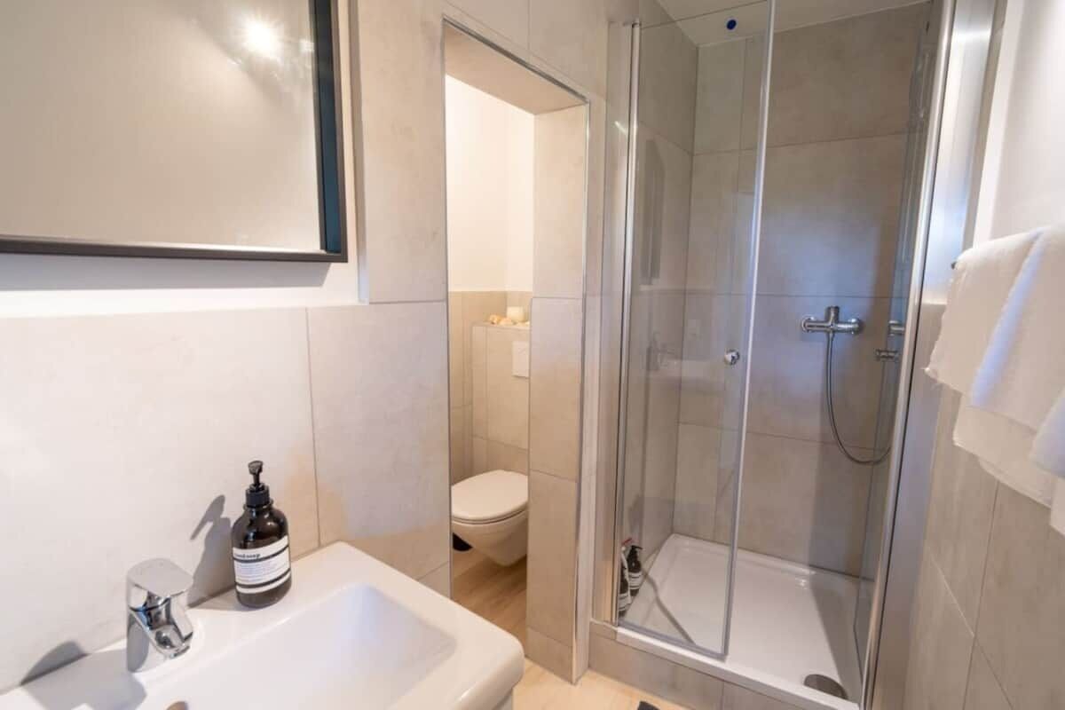 Ein modernes Badezimmer mit einer ein Glasfachwerk umgebenen Dusche, einem weißen Waschbecken und einer Toilette, die durch eine offene Tür sichtbar ist.