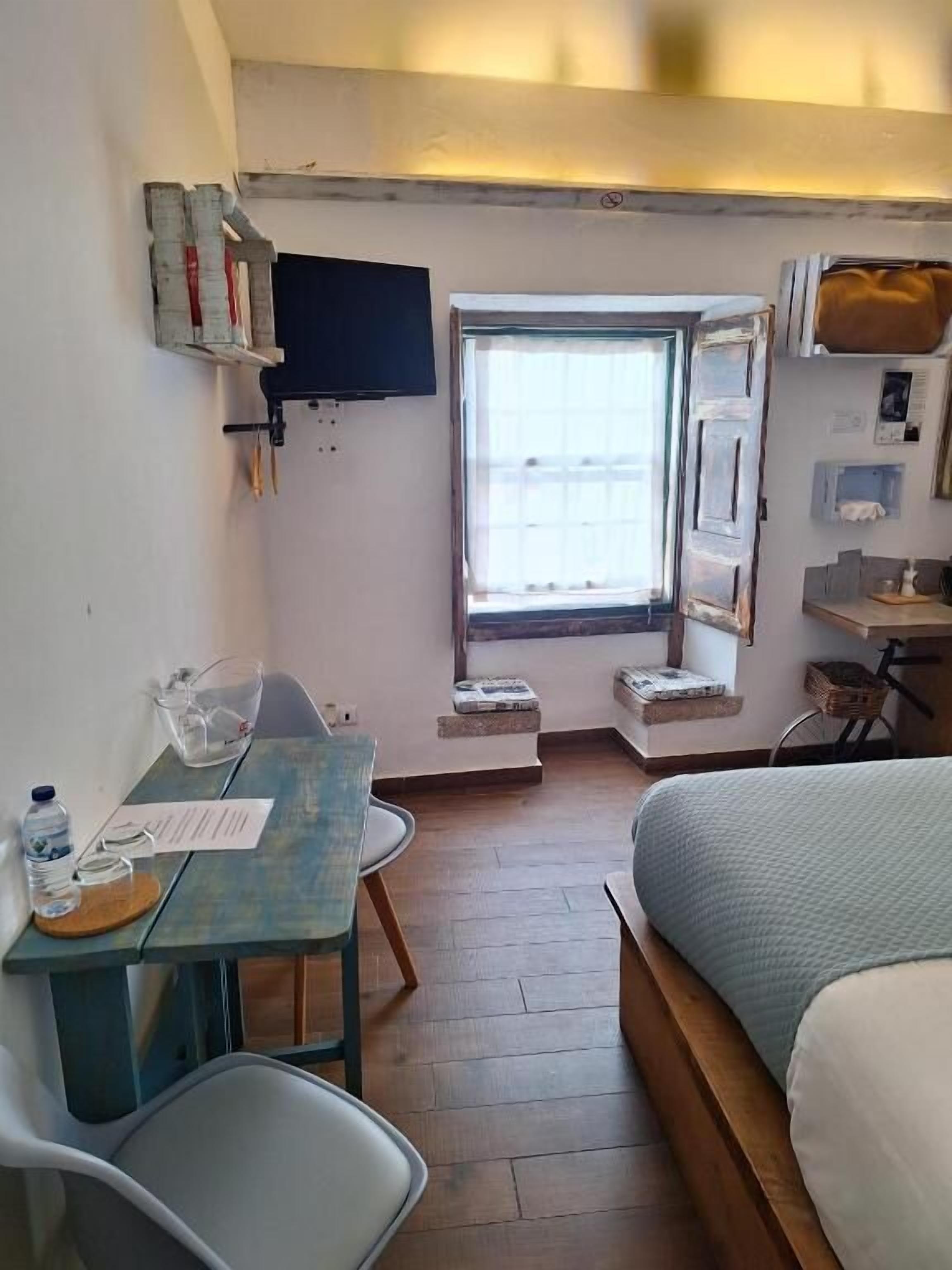 A compact room with a bed, a small table, a chair, a window, and a television mounted on the wall.
