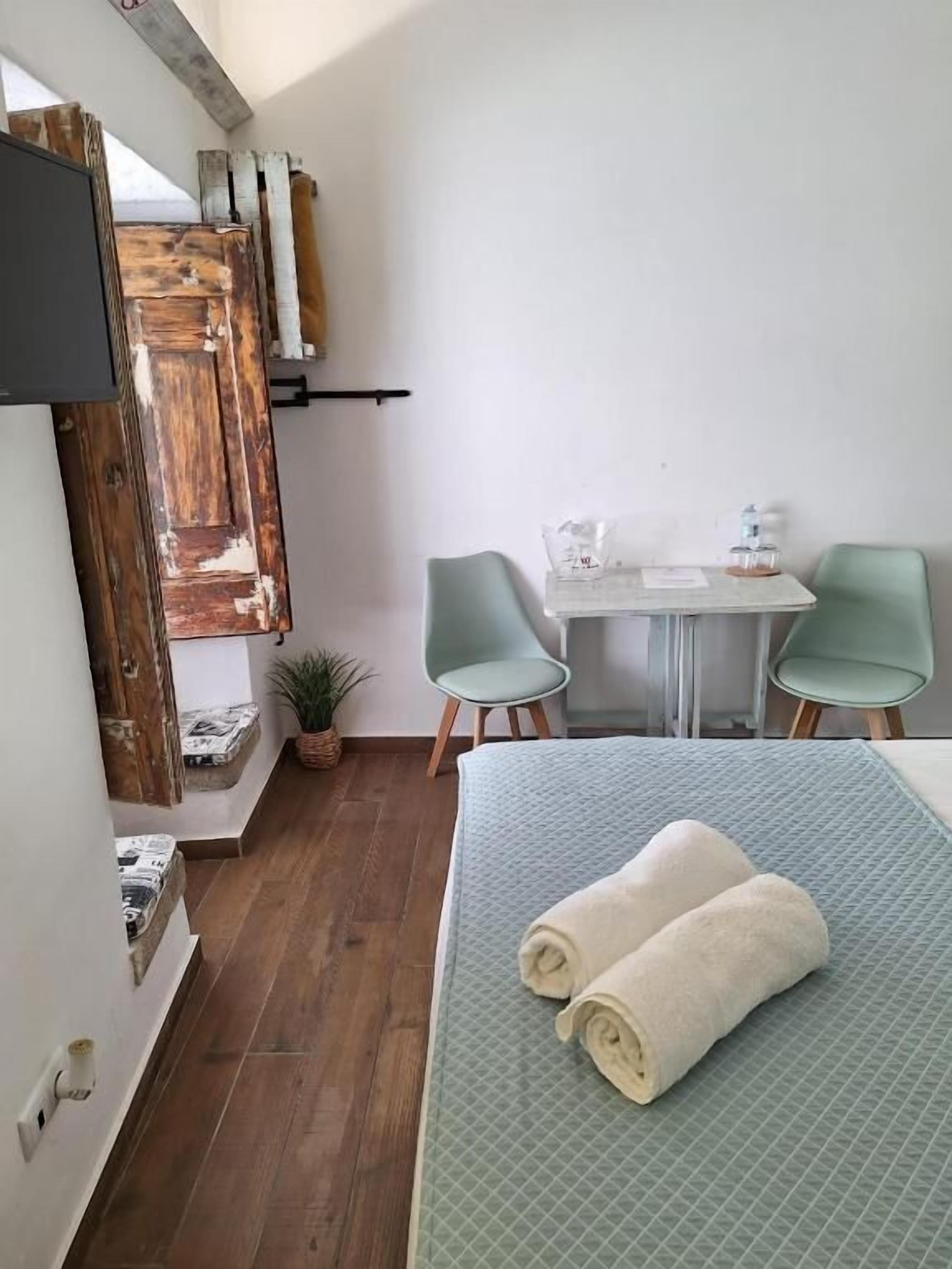 A small, cozy room with a bed, two chairs, a table, and a TV mounted on the wall.