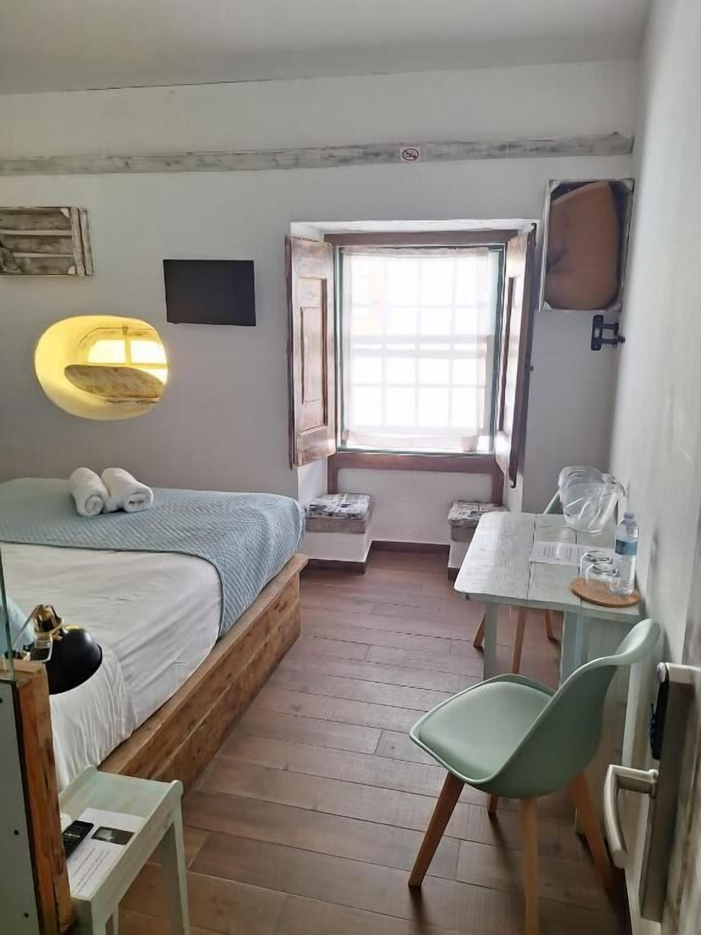 Une petite pièce comprenant un lit, un bureau, une chaise, une fenêtre et un luminaire fixé au mur.