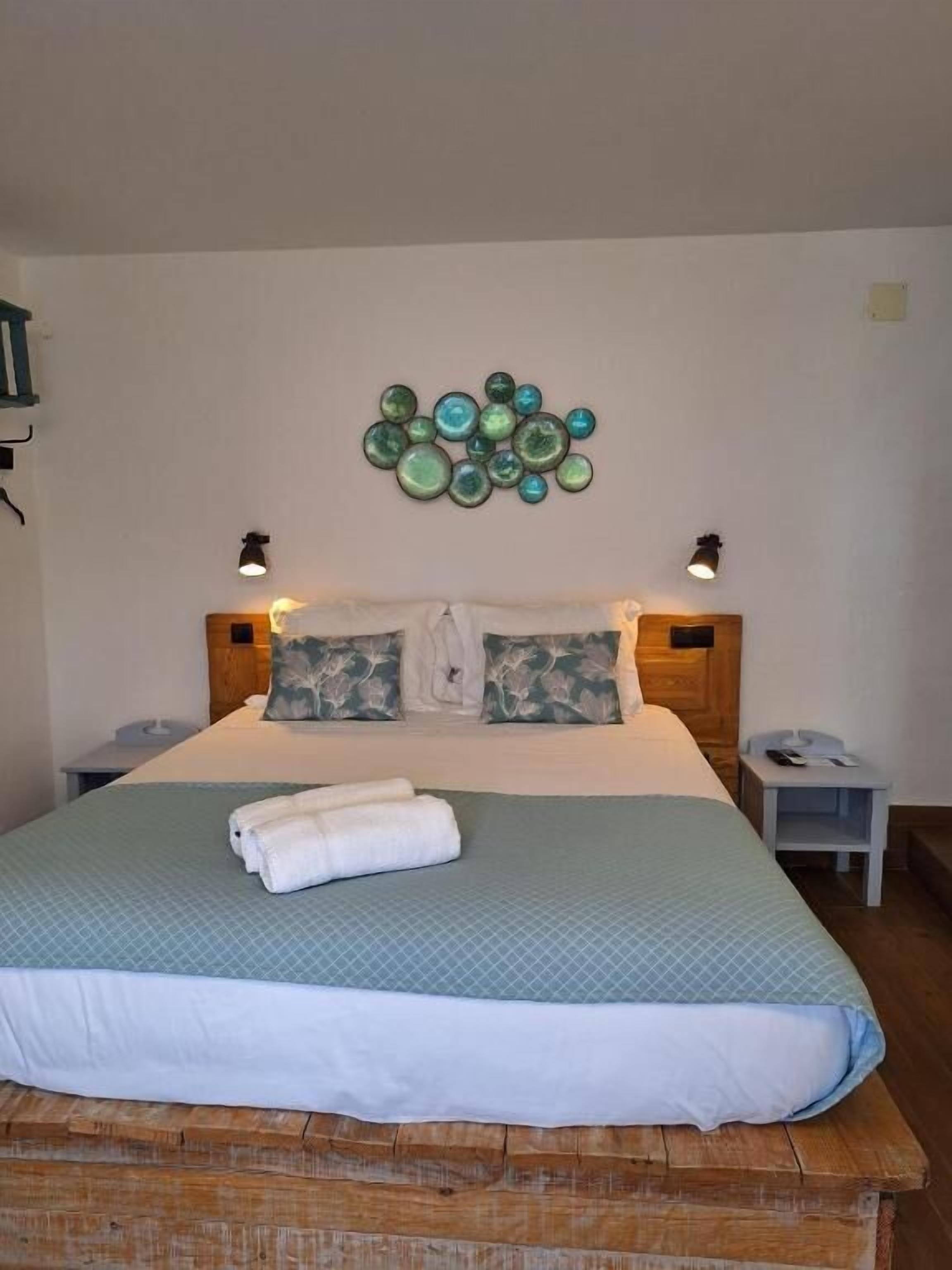A neatly made bed with a blue quilt, two pillows, and a folded towel. There are two bedside tables with lamps and a wall decoration above the bed.
