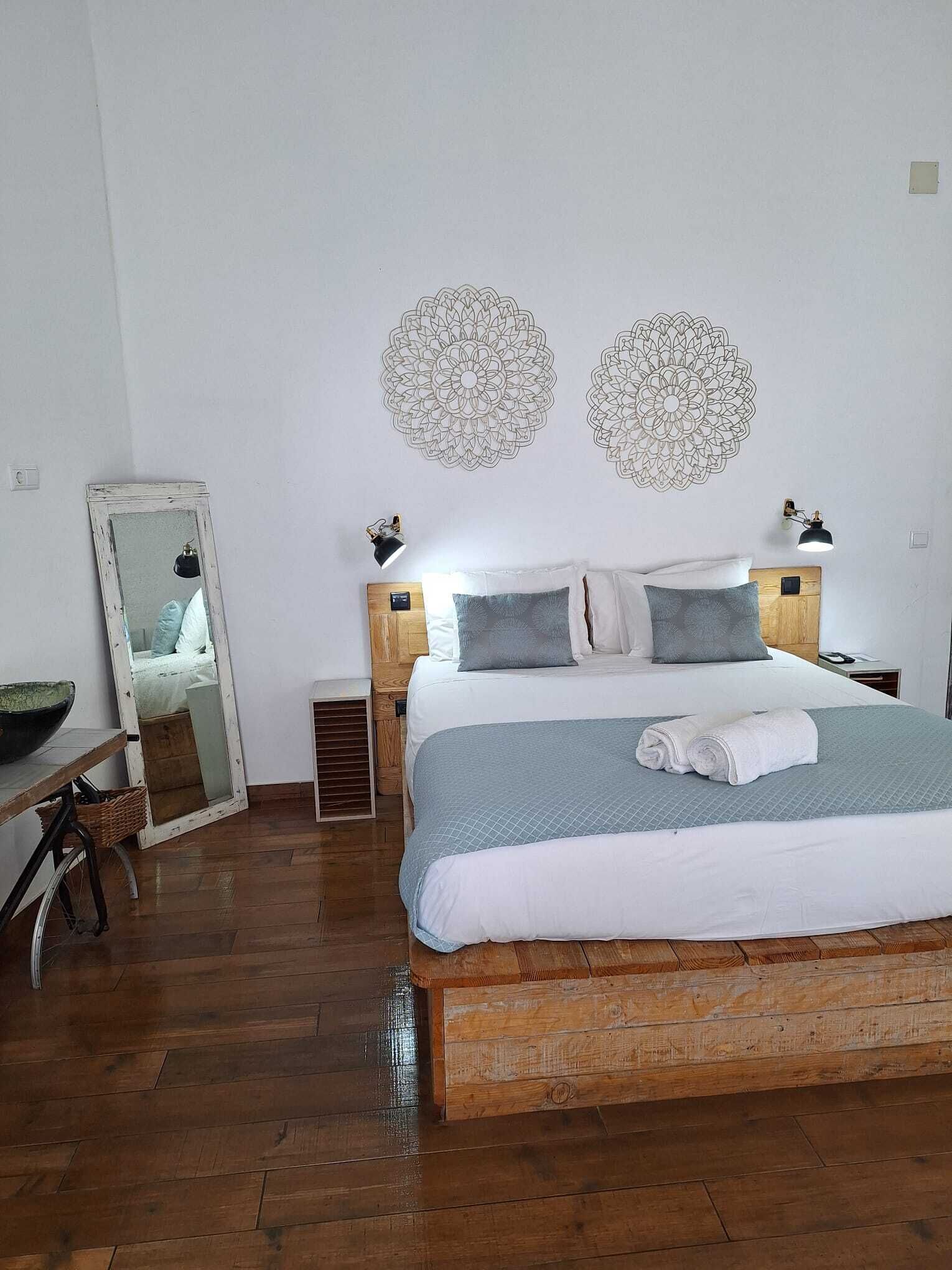 Un dormitorio con cama de madera, ropa de cama blanca, dos lámparas fijadas a la pared, un espejo grande y una mesita de noche de madera con una lámpara.