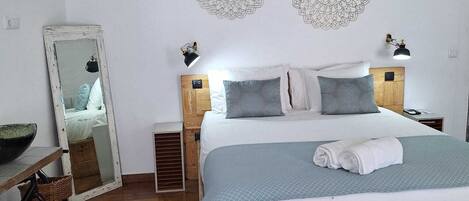 Un dormitorio con cama de madera, ropa de cama blanca, dos lámparas fijadas a la pared, un espejo grande y una mesita de noche de madera con una lámpara.