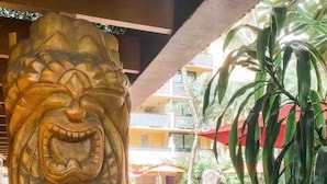 Eine hölzerne Löwenstatue mit markantem Gesicht, auf einem Steinsockel platziert. Im Hintergrund ein Poolbereich mit Liegestühlen und Sonnenschirmen, umgeben von Blumentöpfen.