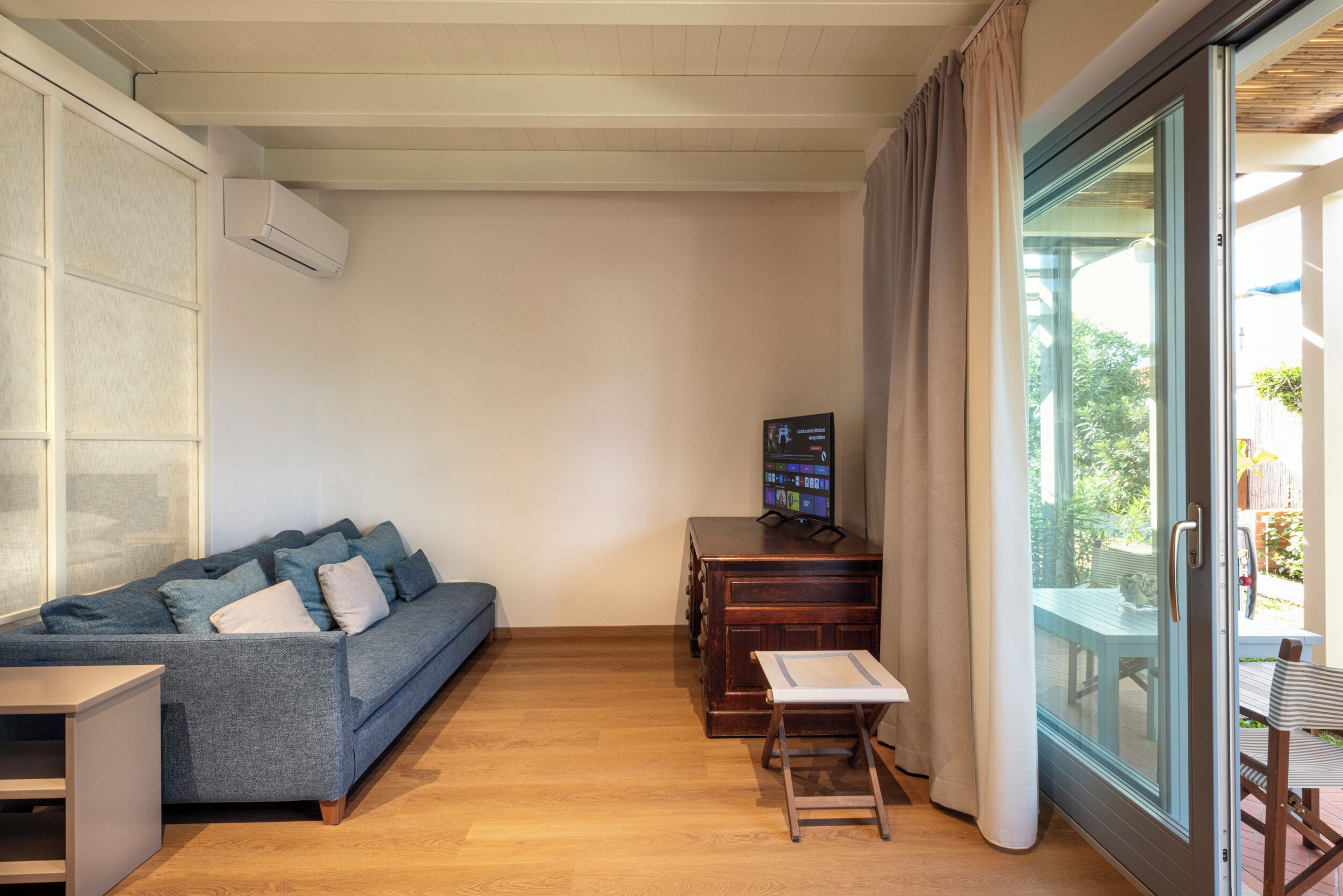 Ein Wohnzimmer mit einer Couch, einem Fernseher auf einer Holzkommode und einer Glastür, die zu einem Außenbereich führt.