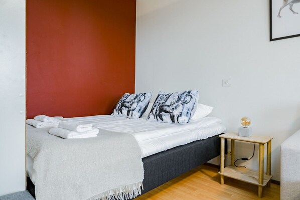 A neatly made bed with white linens and a grey throw blanket, a bedside table with a lamp, and a red accent wall.