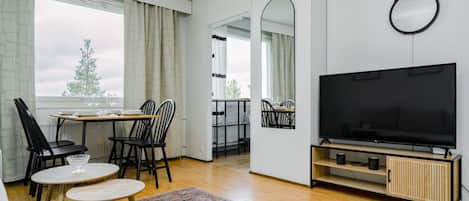 Un salon moderne doté d’un téléviseur à écran plat, d’un coin repas avec des chaises et une table, et d’une grande fenêtre agrémentée de rideaux.