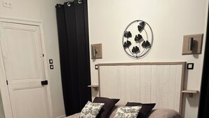 Quarto com cama, cobertor preto e bege, e duas almofadas. Há uma cabeceira de madeira, um enfeite fixado na parede e uma porta branca.