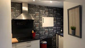 A modern kitchen with a black backsplash featuring coffee-related text, a stainless steel range hood, and a red trash bin. - Holiday Home "Gîte Les Aubennes" with Private Terrace, Garden & Wi-Fi (Fromelennes)