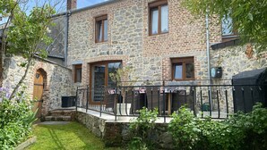 A stone building with a wooden door, a balcony with wicker furniture, and a garden. - Holiday Home "Gîte Les Aubennes" with Private Terrace, Garden & Wi-Fi (Fromelennes)