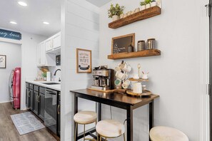 Uma cozinha moderna com armários brancos, bancada preta e banquetas. Há prateleiras com plantas e utensílios de cozinha, e uma máquina de café no balcão.