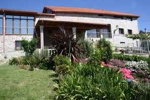 A stone building with a balcony, surrounded by a garden with various plants and flowers.