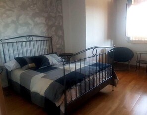 A bed with a metal frame, a small round table, and a chair in a room with patterned wallpaper and a window.