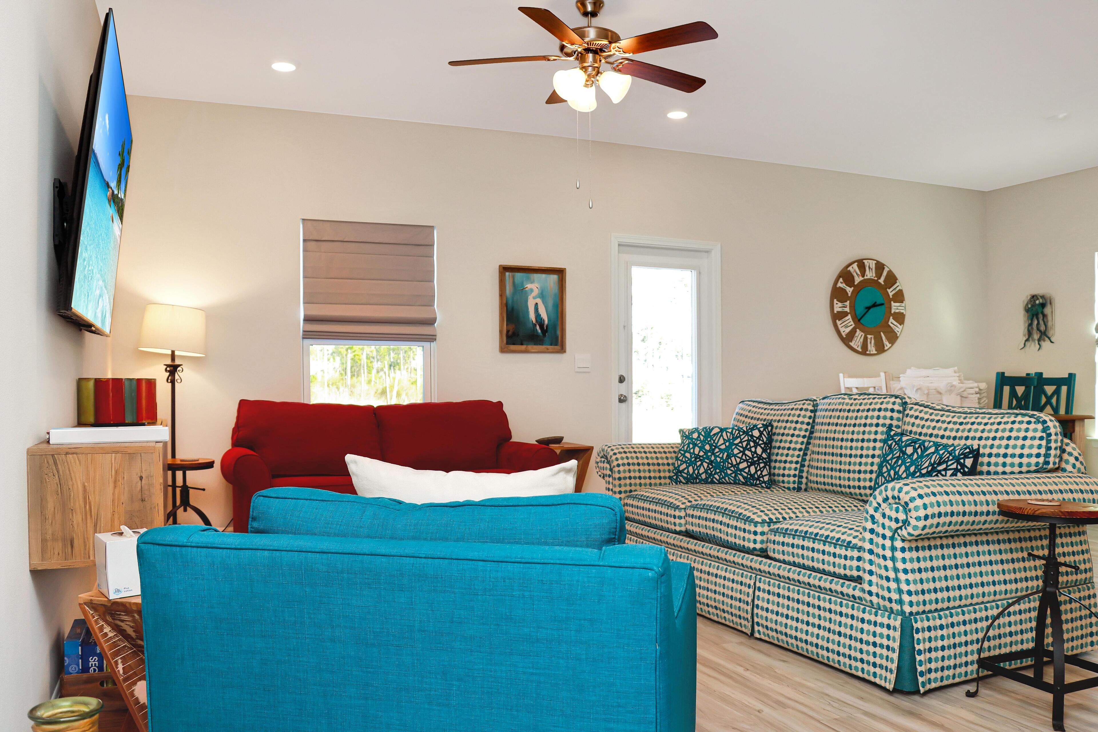 A living room with a blue sofa, a red sofa, and a patterned sofa. There is a ceiling fan, a clock, and a TV mounted on the wall.
