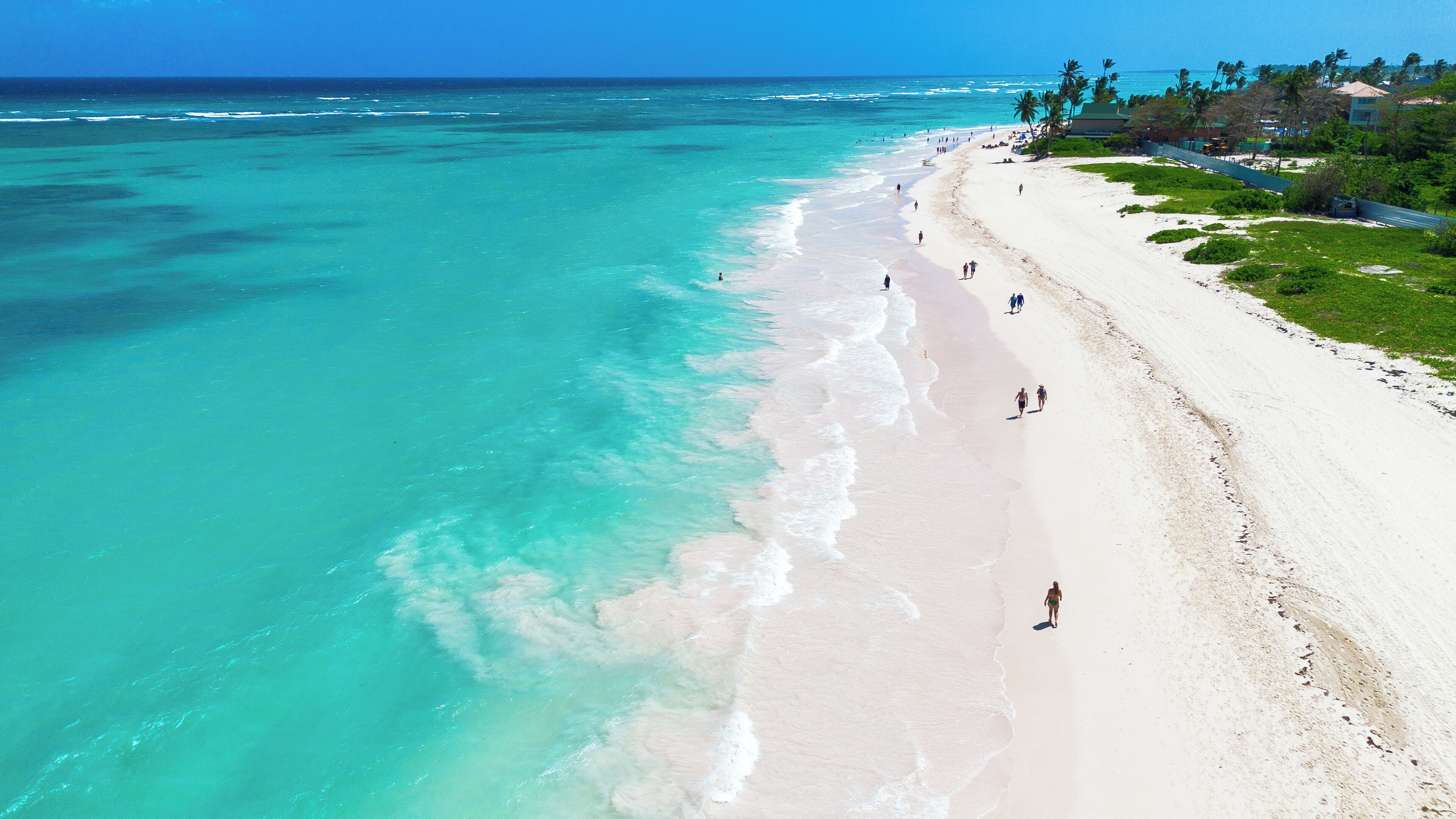 A beach with clear turquoise water, white sand, and a few people walking along the shore.