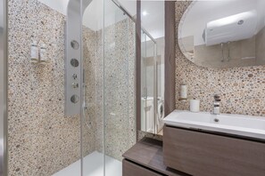 Ein modernes Badezimmer mit einer Wand aus Kieselmoos, einer Glaskabinentrennwand für die Dusche, einem weißen Waschbecken und einem Spiegel mit Holzrahmen.