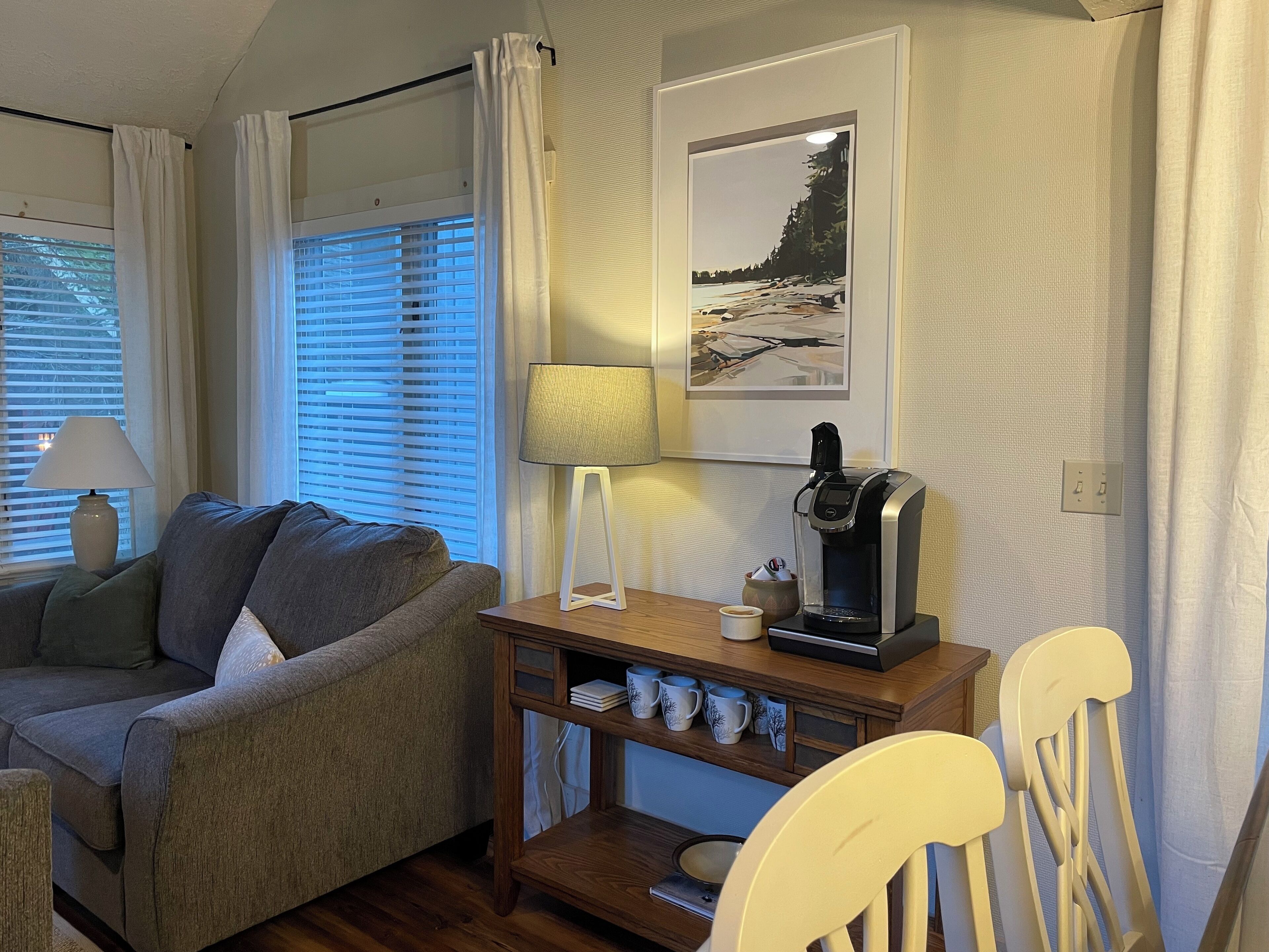 A cozy room with a sofa, a coffee table with a coffee maker, a lamp, and a framed picture on the wall.
