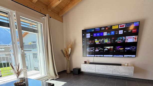 Una sala de estar moderna con un televisor de pantalla plana grande montado en la pared, una mesa de centro de mármol negro, un tapete beige con textura y vistas a un balcón con una montaña al fondo.
