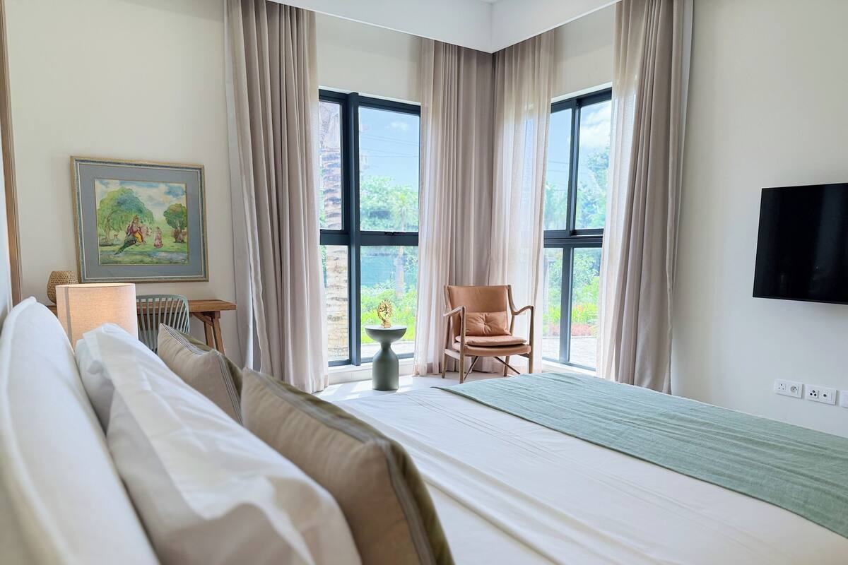 A hotel room with a bed, a chair, a table, a painting, and a TV.