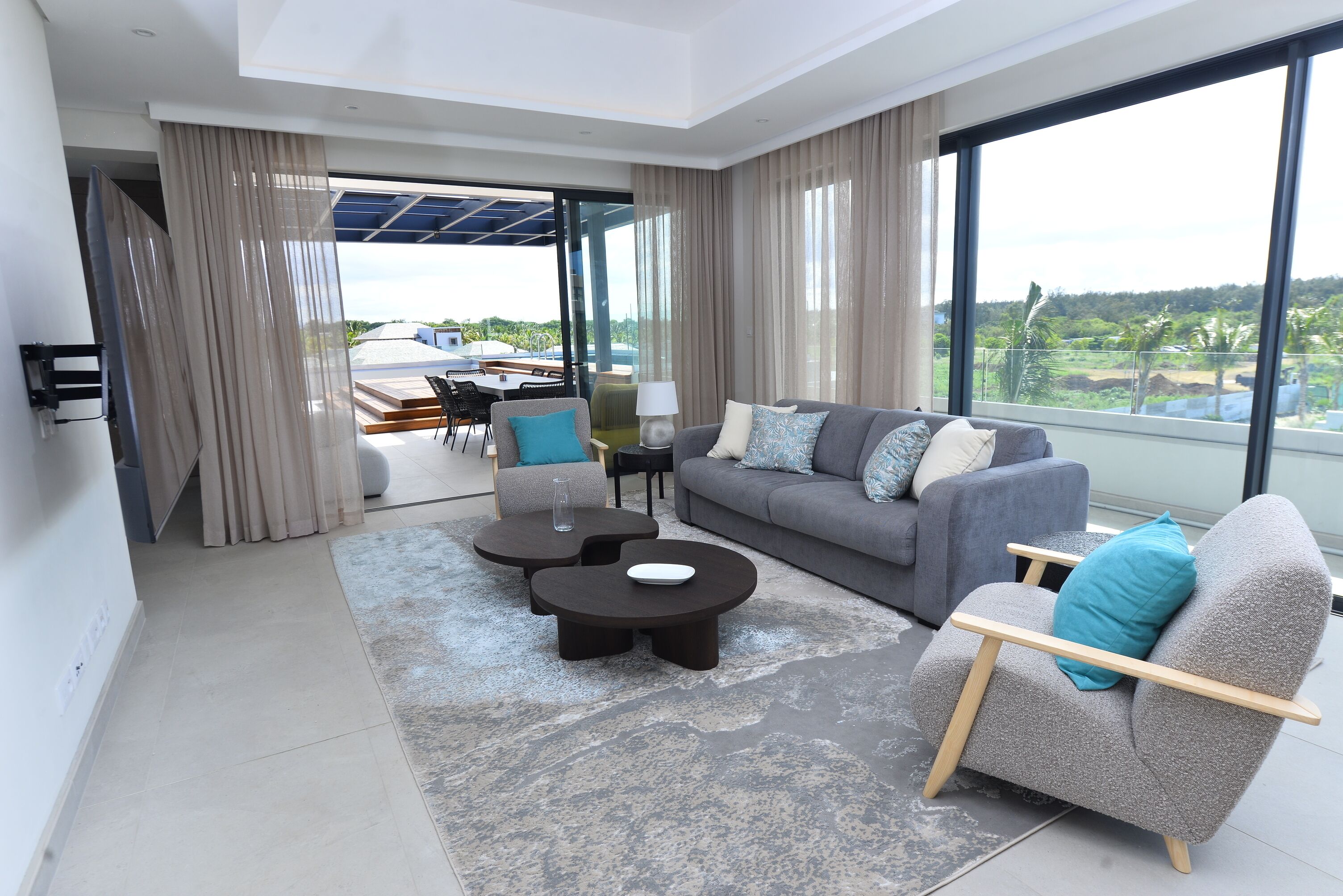 A modern living room with a grey sofa, two round coffee tables, and a grey armchair. There is a large window with a view of greenery and a patio area with outdoor furniture.