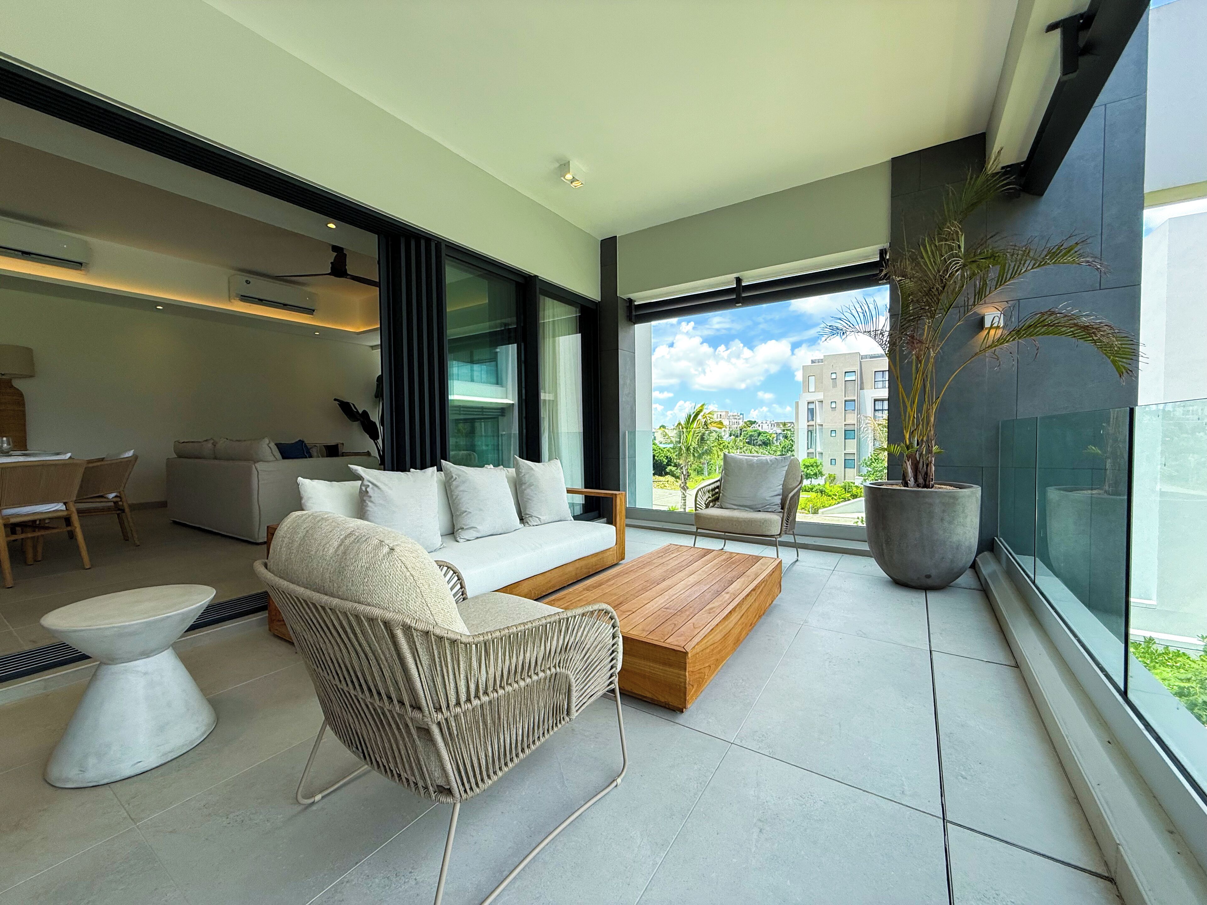 A modern living room with a sofa, coffee table, and chairs, a dining area with a table and chairs, and a large window with a view of buildings and greenery.