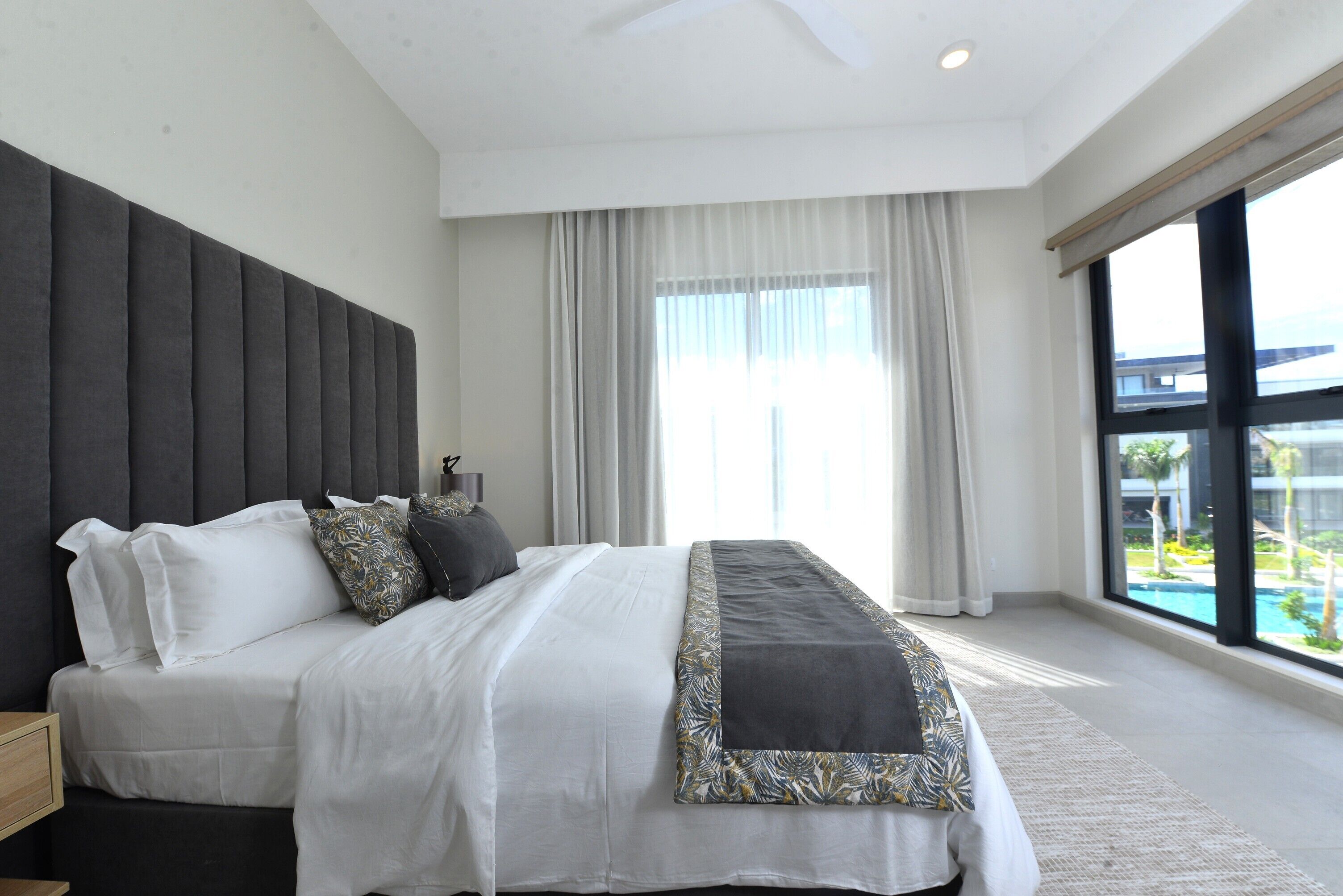 A modern bedroom with a large bed, a headboard, and a view of a swimming pool through large windows.