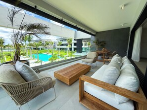 A modern living room with a sofa, a wooden coffee table, and a wicker chair. There is a view of a swimming pool and buildings in the background.