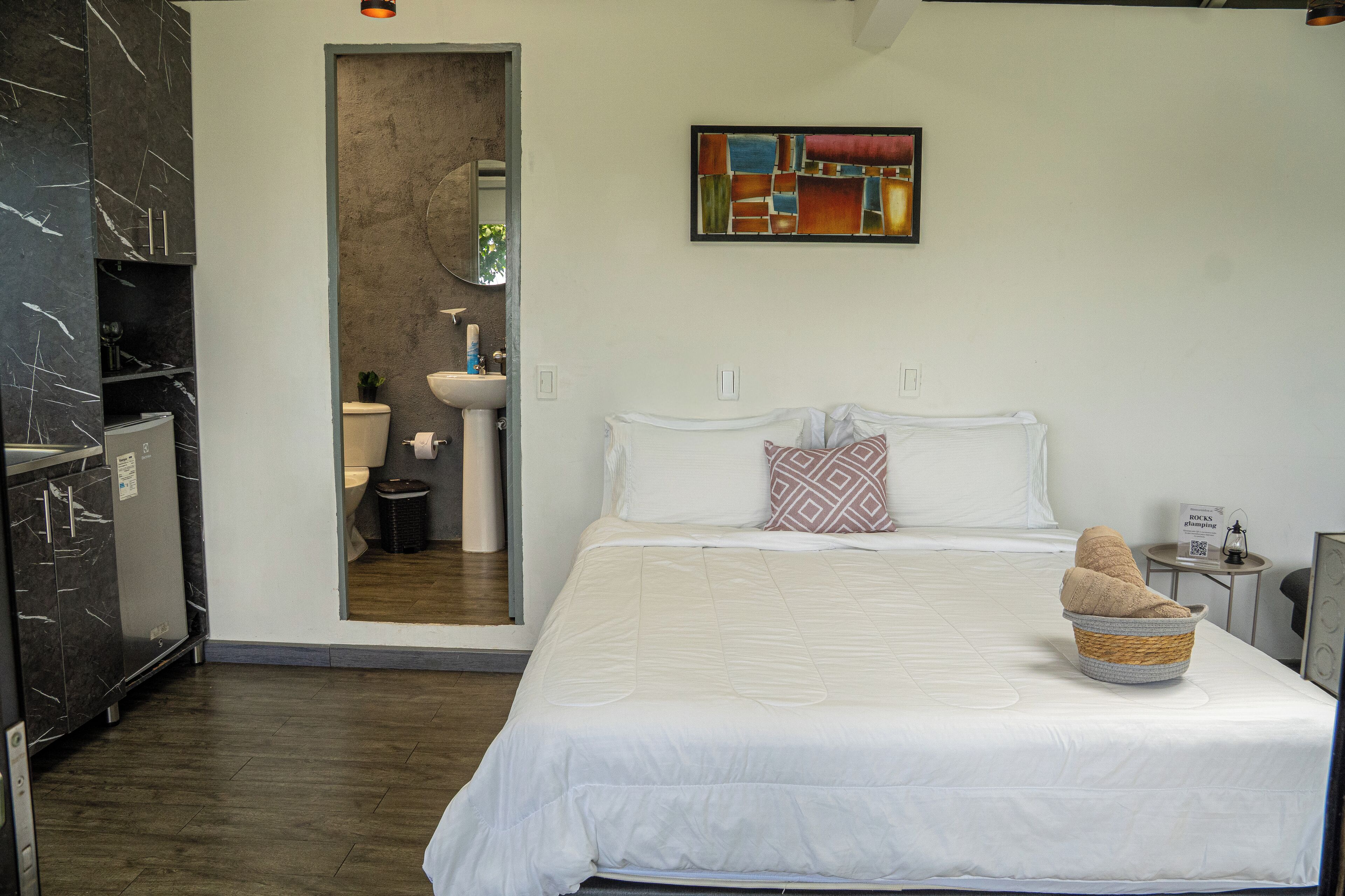 A modern bedroom with a large bed, a small bedside table, a wall-mounted painting, and a bathroom visible through an open door.